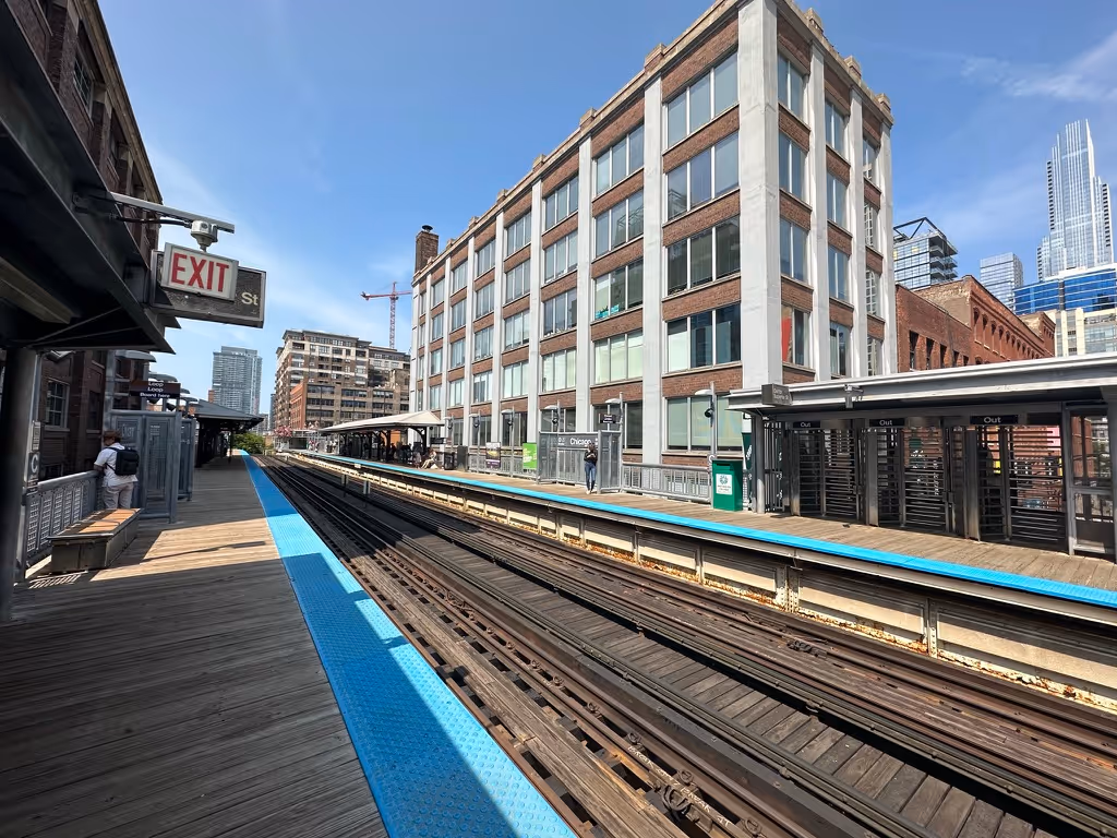 Empty elevated train platform with tracks running through a cityscape of mid-rise and high-rise buildings under a clear blue sky.