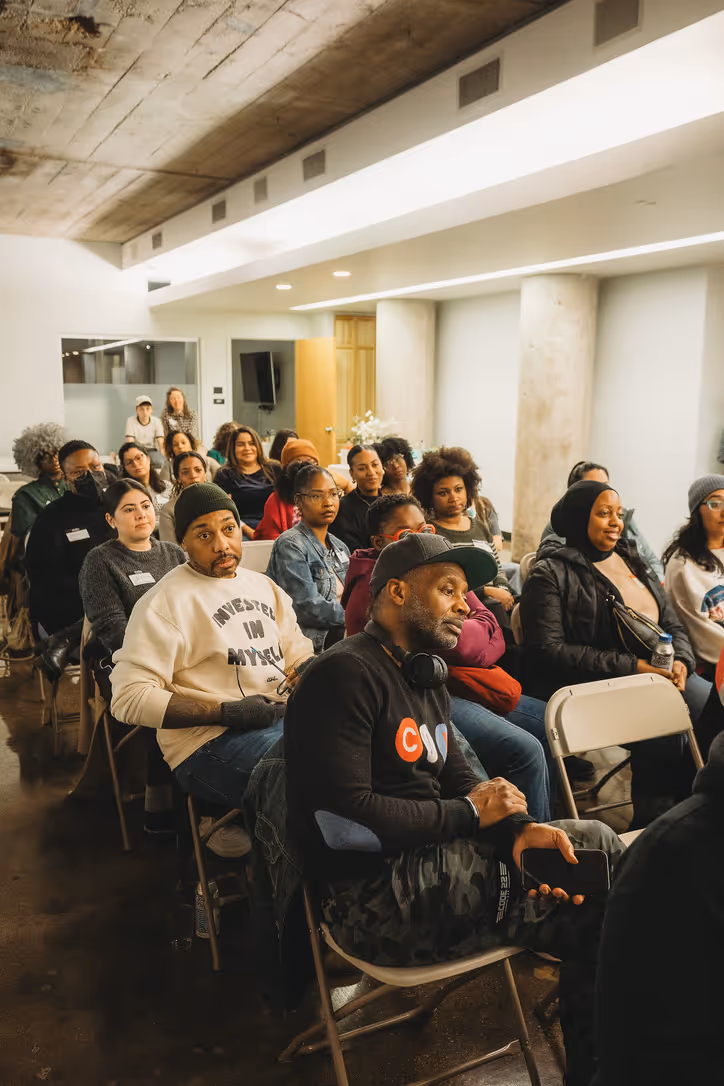 A diverse group of people attentively seated in rows inside a modern room with concrete pillars and ceiling.