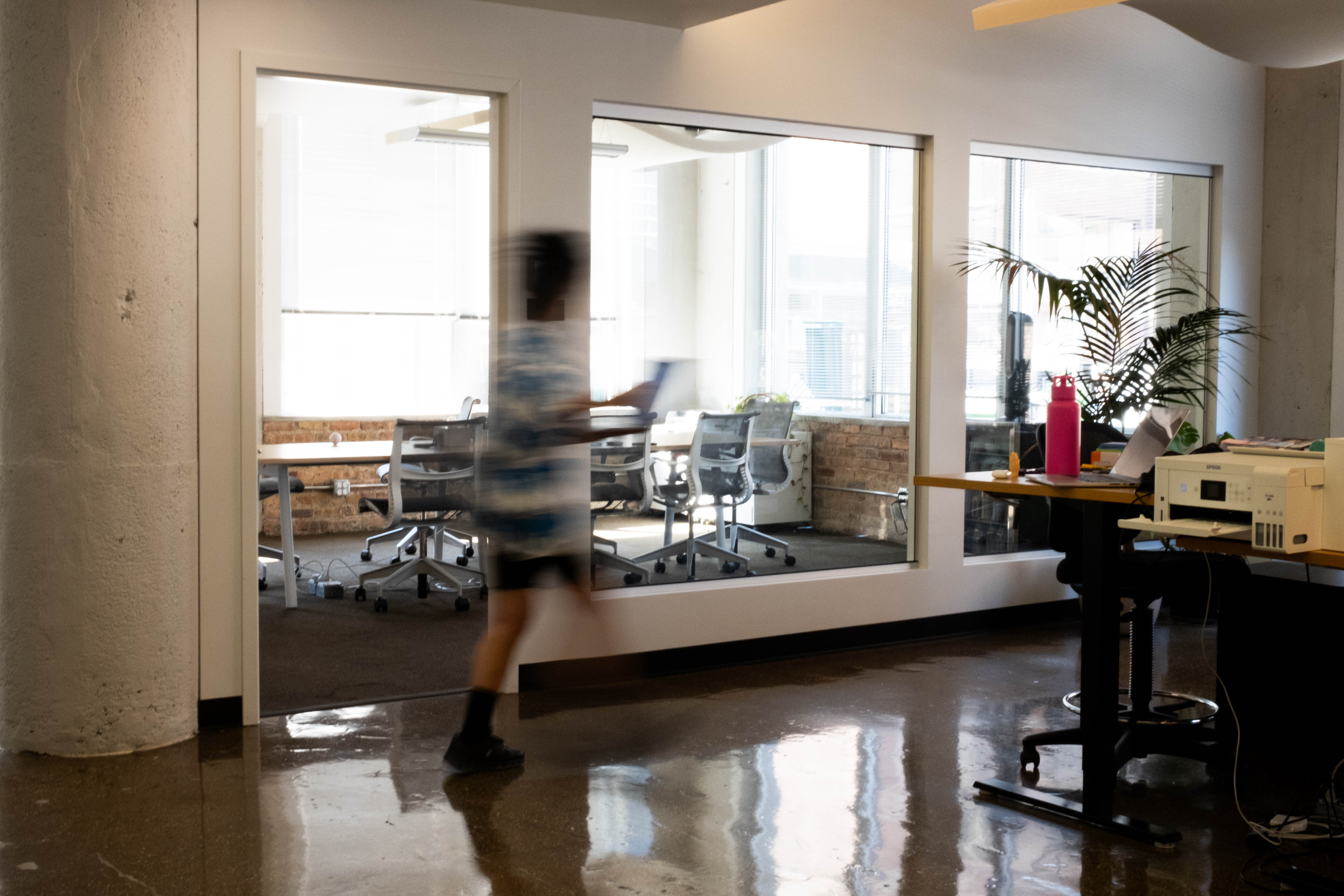 Blurred person walking through a modern office space with glass walls, office chairs, and a desk with a printer and pink water bottle.