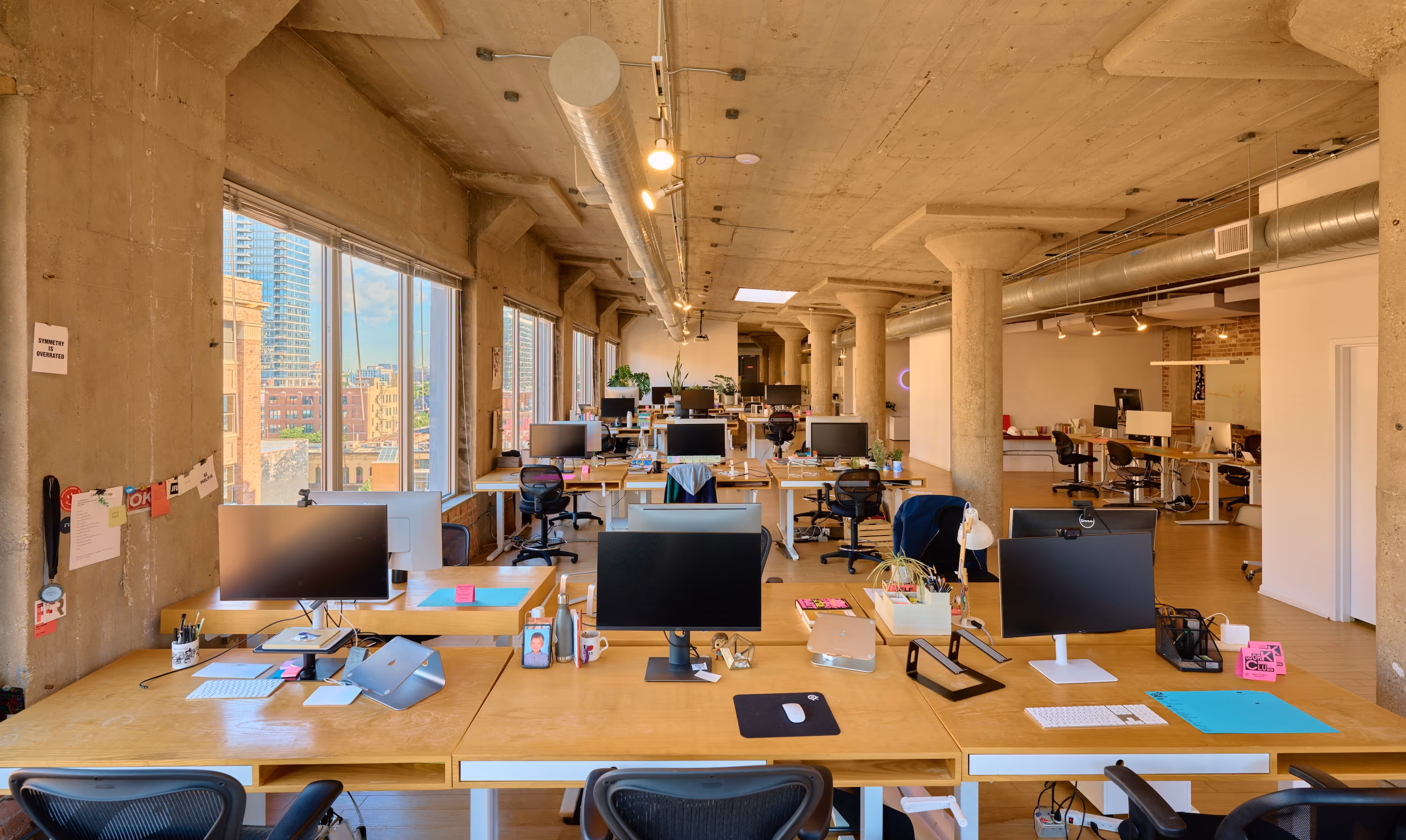 Modern open office space with concrete pillars, large windows, multiple desks with computer monitors, and three potted plants on a wooden divider.