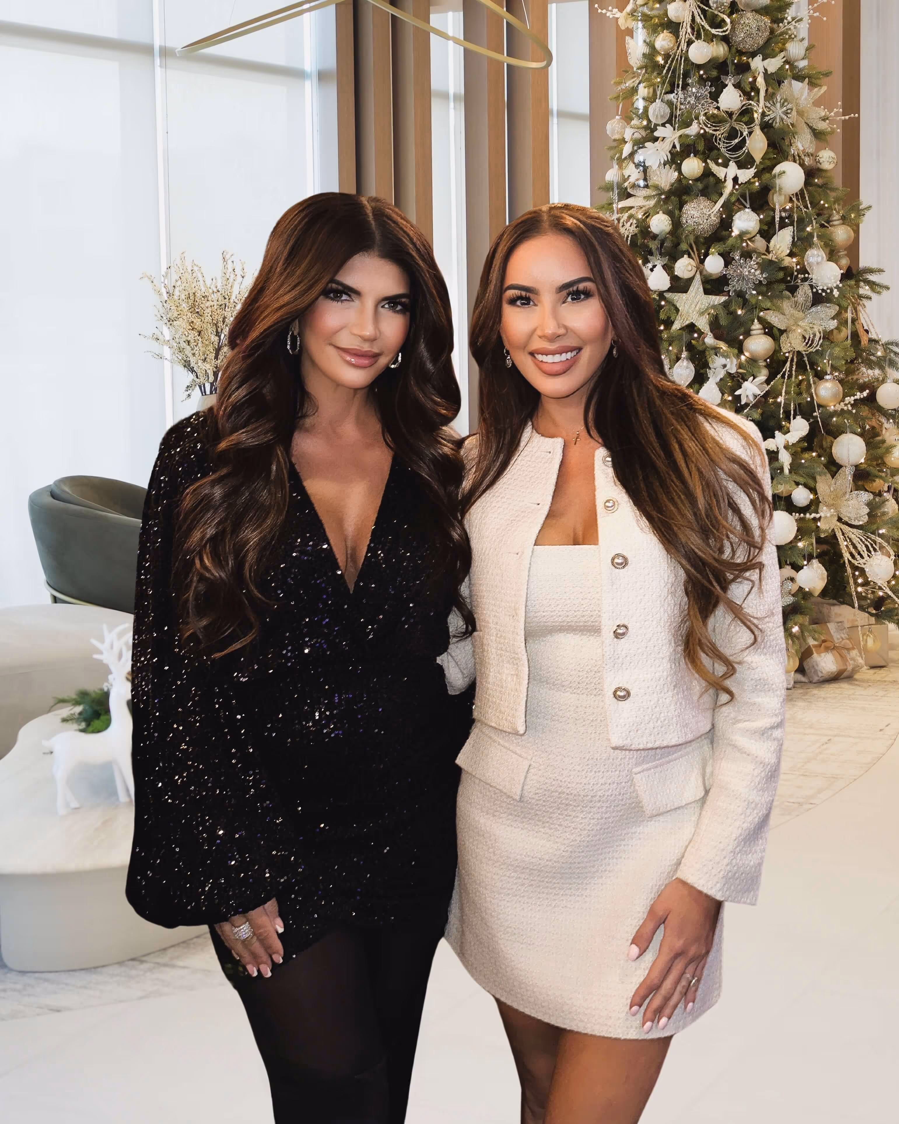 Two women standing together smiling, one in a black sequined dress and the other in a white textured dress with a matching jacket, with a decorated Christmas tree in the background.