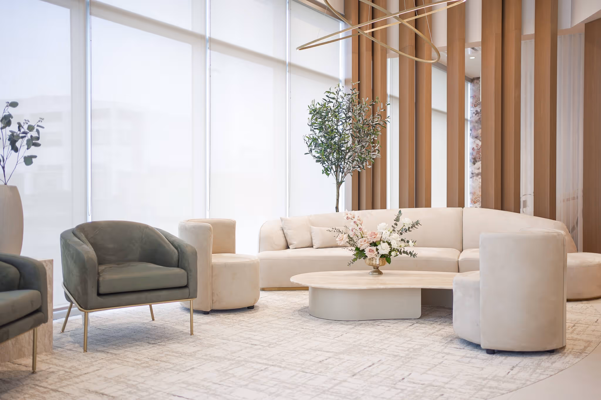 Modern living room with beige curved sofa, two armchairs, round wooden coffee table with flowers, and tall plant near large windows with blinds.