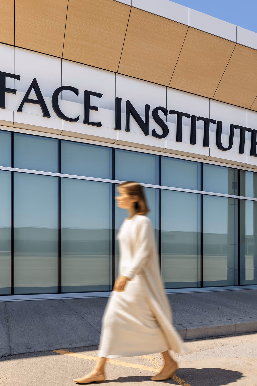 Blurred woman in beige dress walking past building with large glass windows and sign reading 'FACE INSTITUTE'.