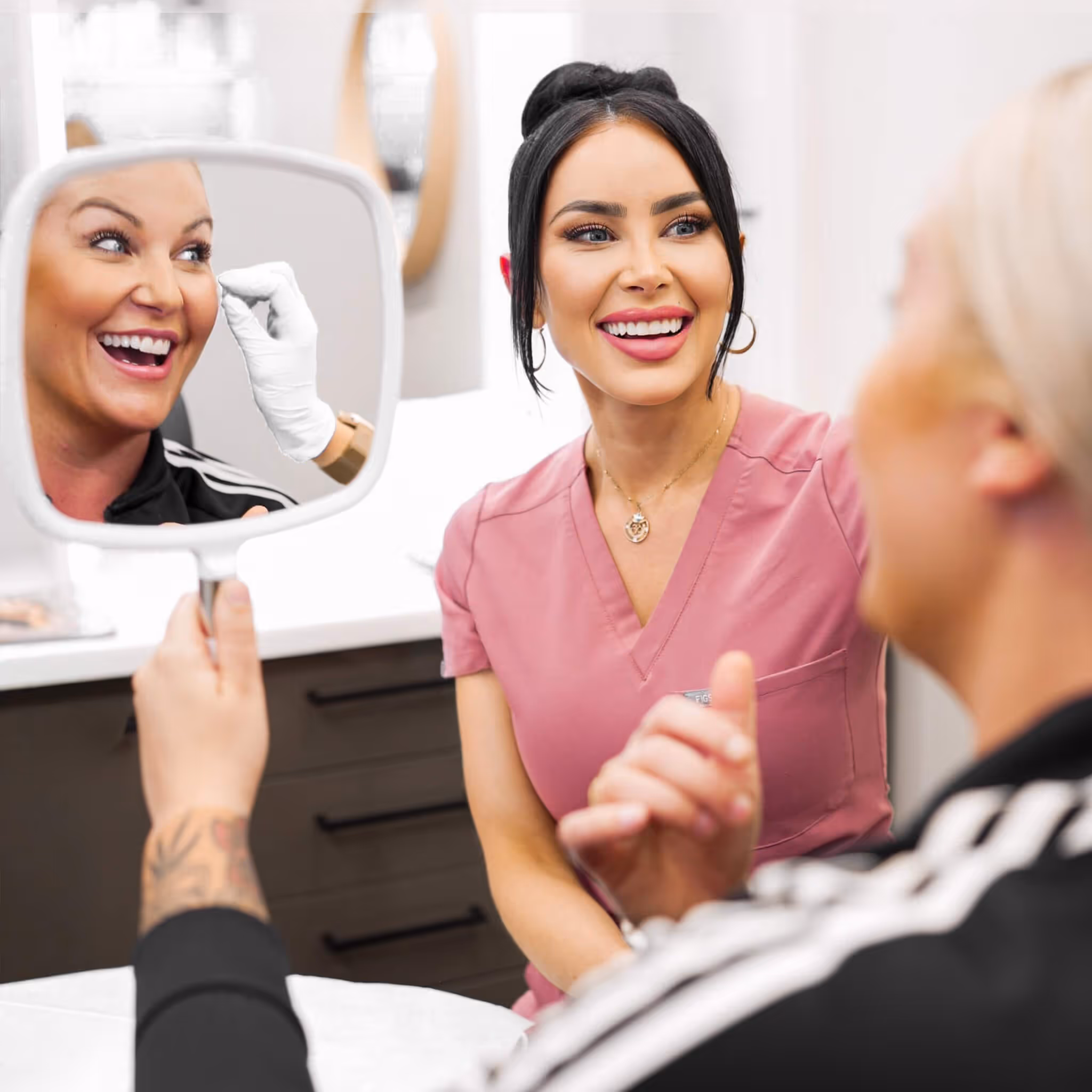 Woman in pink scrubs smiling at a client holding a mirror who is happy after a cosmetic procedure.