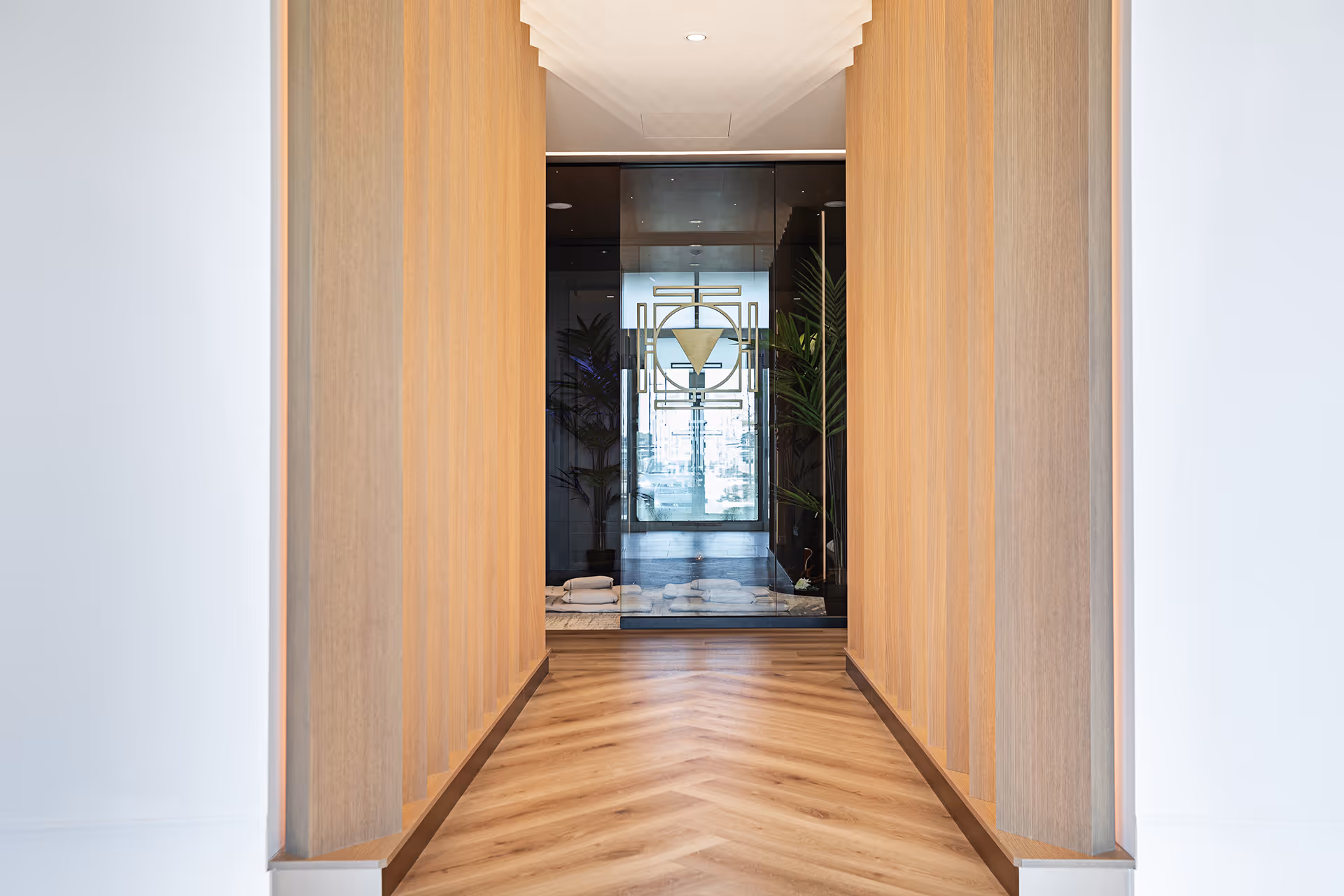 Modern hallway with wooden panel walls and chevron wood floor leading to a glass door decorated with a geometric pattern and plants inside.