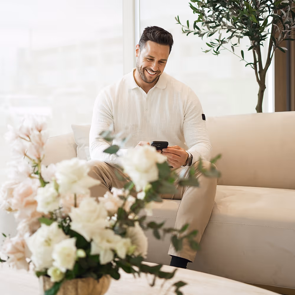 Smiling man in a white sweater sitting on a beige couch and using a smartphone with a floral arrangement in the foreground.