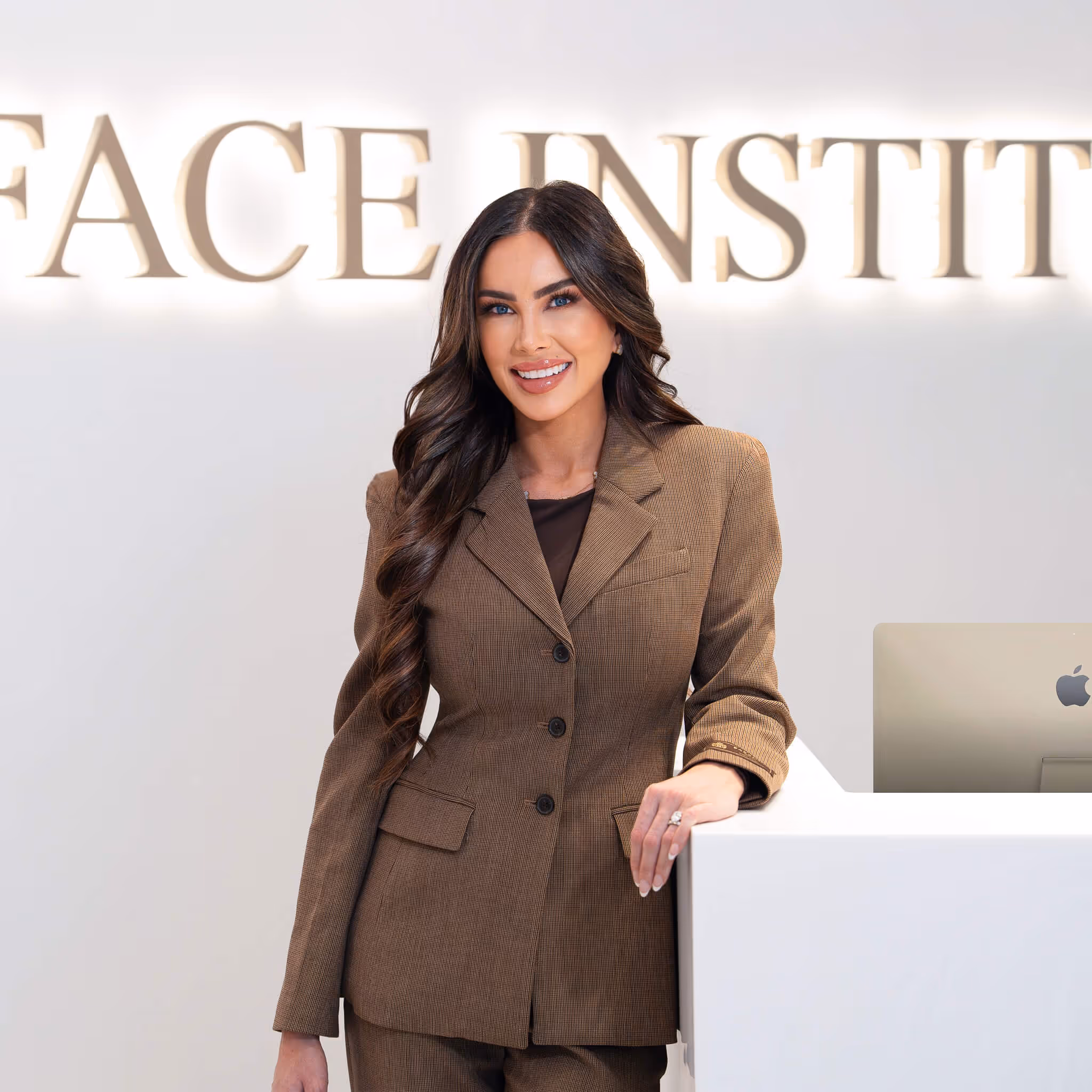 Smiling woman with long dark hair in a brown suit standing at a white reception desk with 'Face Institute' sign behind her.
