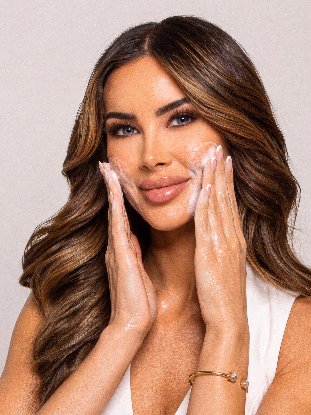 Woman with long hair gently applying foamy cleanser to her cheeks, demonstrating skincare routine.