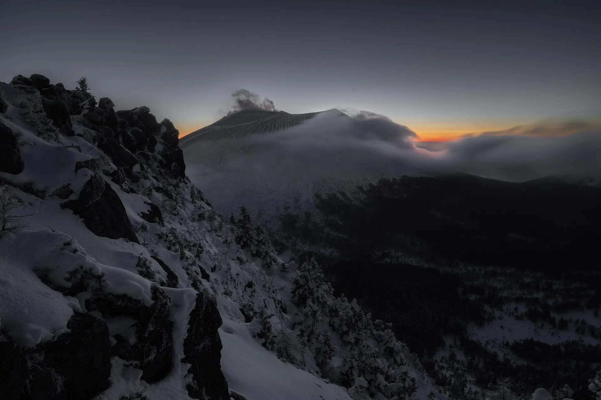 Snow-covered rocky mountain slope with trees at dusk and a mountain peak shrouded in clouds under a darkening sky.