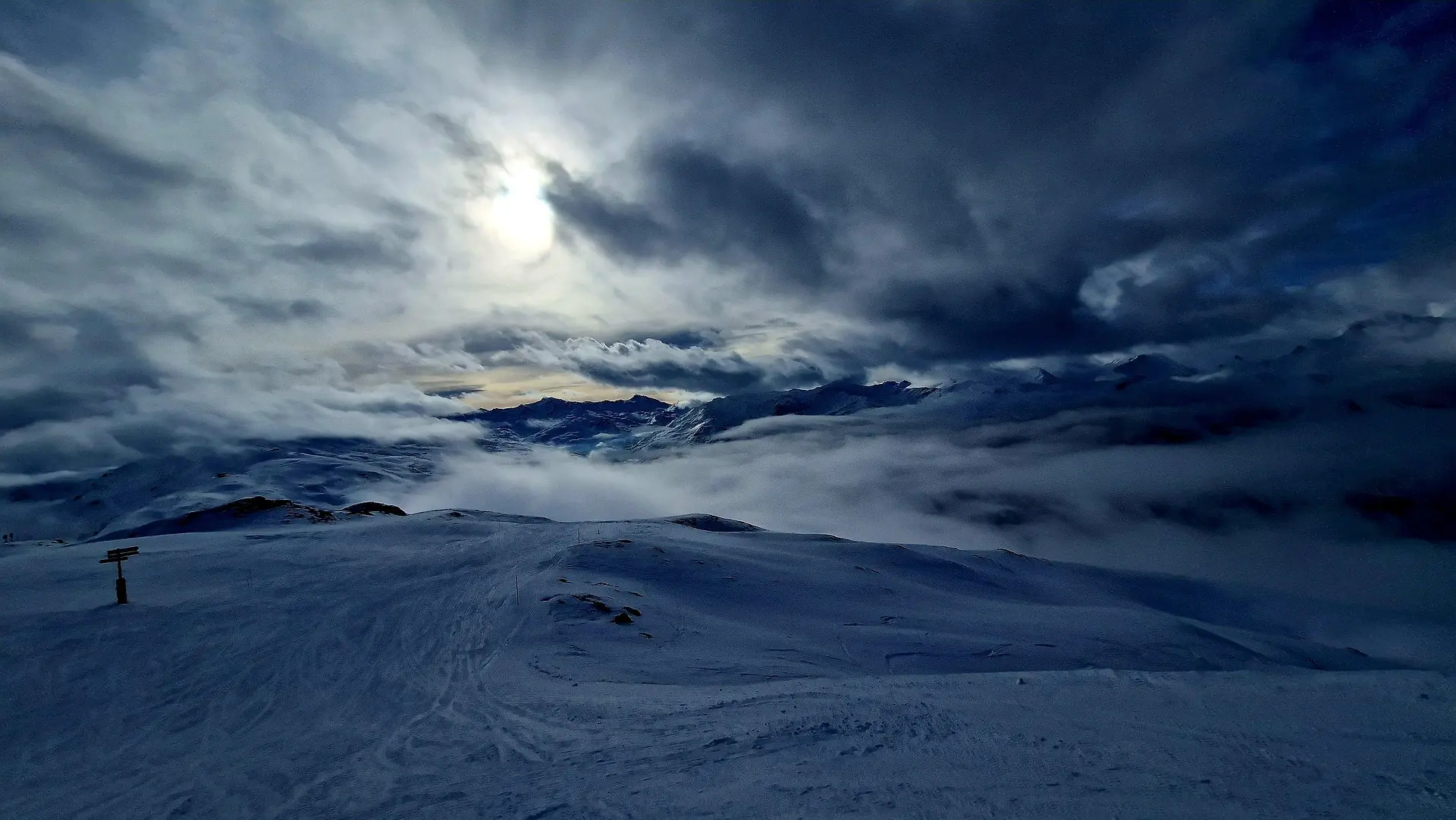 Snow-covered mountain landscape under a cloudy sky with the sun behind clouds at dusk.