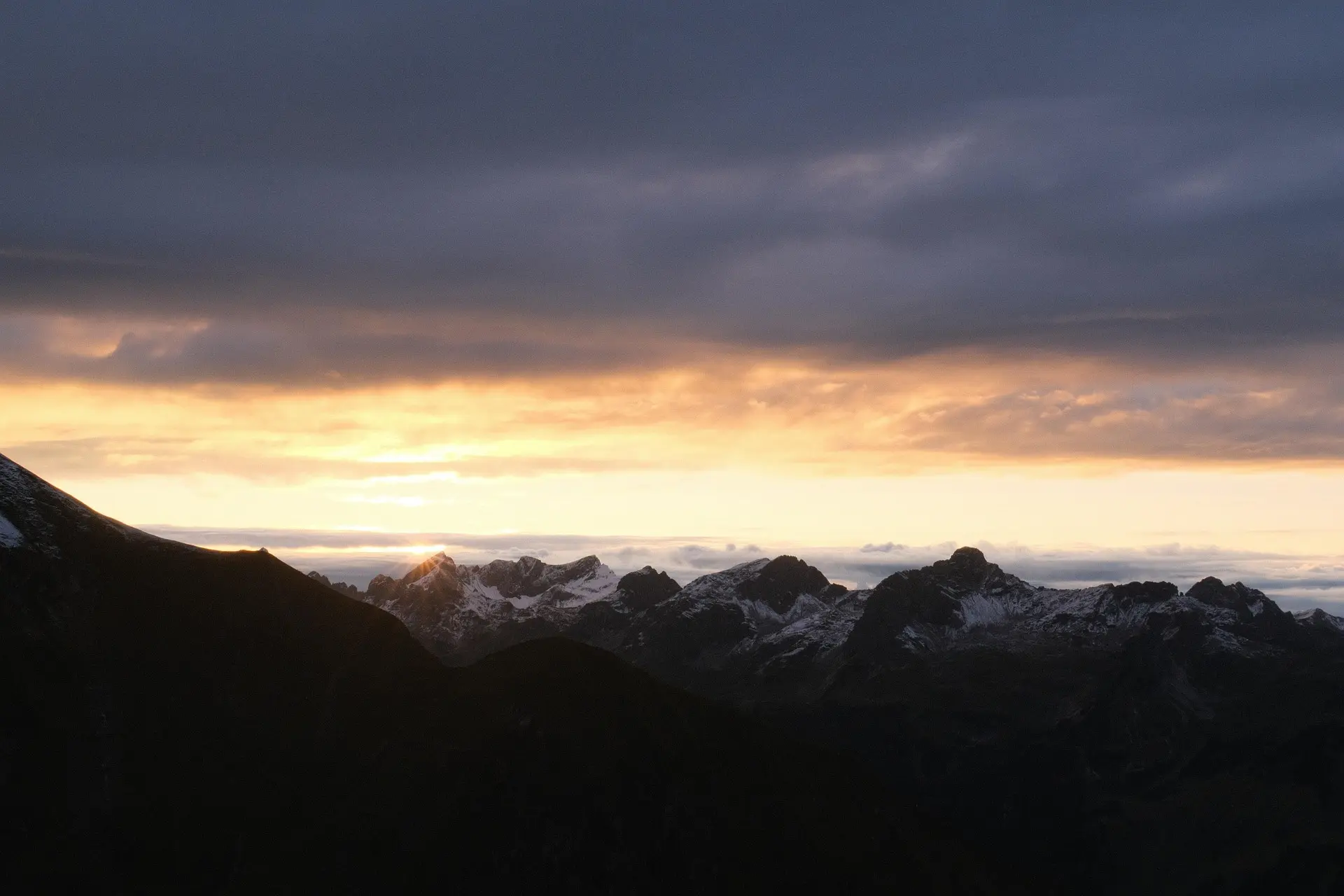 Sunset behind a mountain range with partly snow-covered peaks under a cloudy sky.