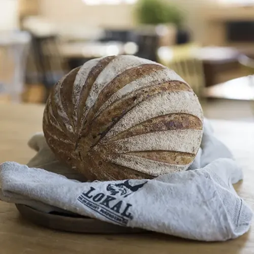 Round loaf of artisan bread with decorative slashes resting on a cloth labeled LOKAL on a wooden table.