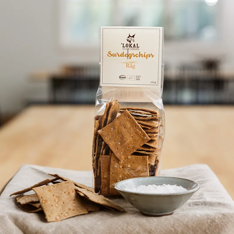 Förpackning med surdegsknäcke av råg, med några knäckebitar och en skål salt på ett träbord med duk.