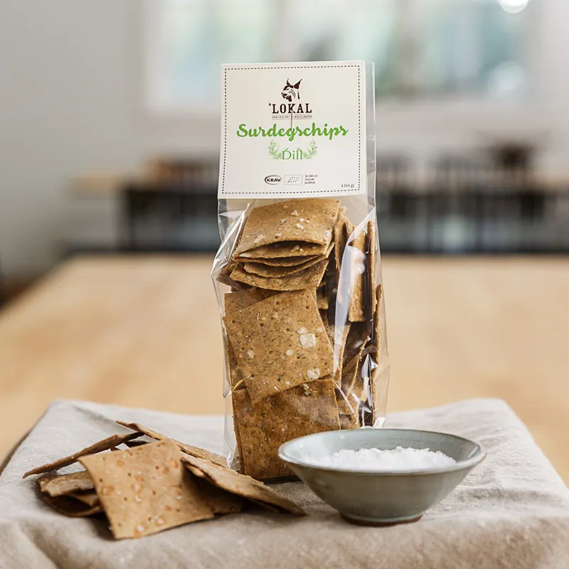 Påse med Lokals surdegschips med dill, med några chips och en skål salt på en duk.