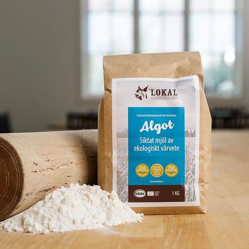 Brun papperspåse med ekologiskt Algot-vetemjöl och en hög mjöl på ett träbord.