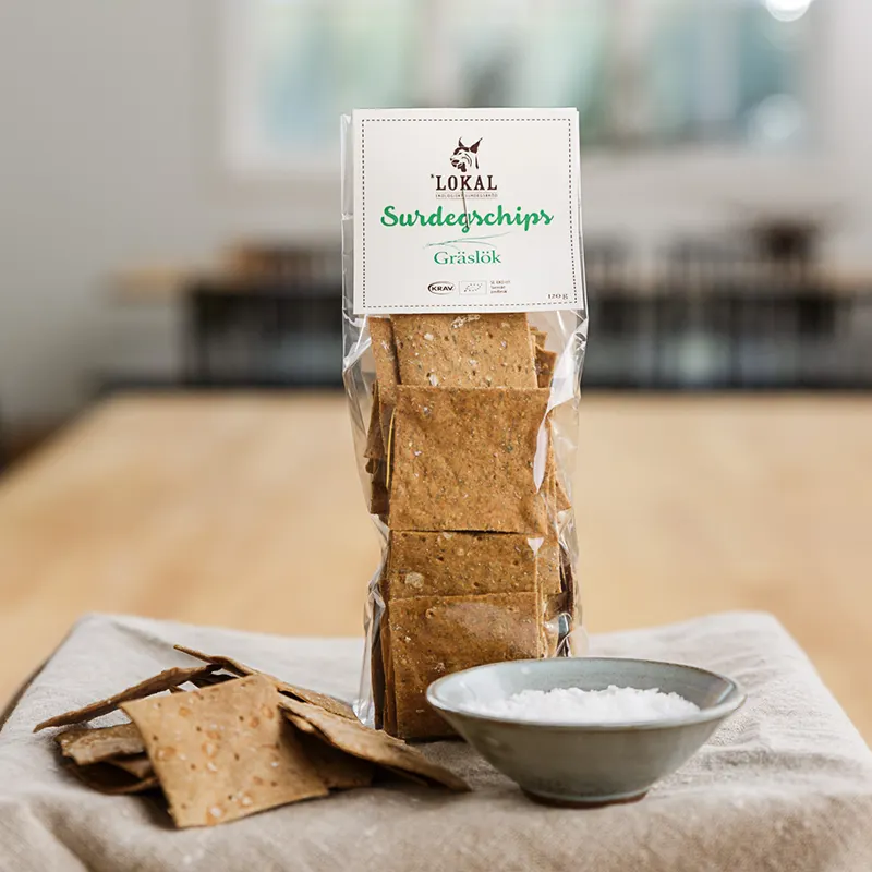 Påse med Lokals surdegschips med gräslökssmak på en duk bredvid en skål med grovsalt.