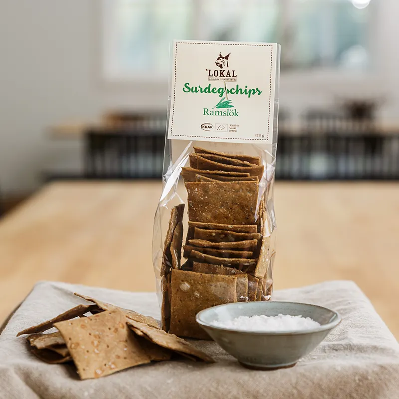 Påse med Lokals surdegschips med ramslök, med krispiga chips och en skål salt på en duk.
