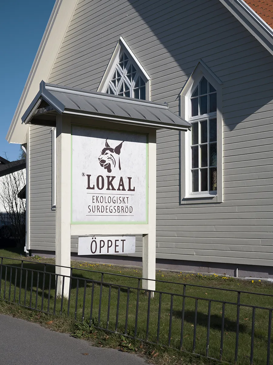 Skylt för Lokal ekologiskt surdegsbröd med texten Öppet utanför en grå träbyggnad.