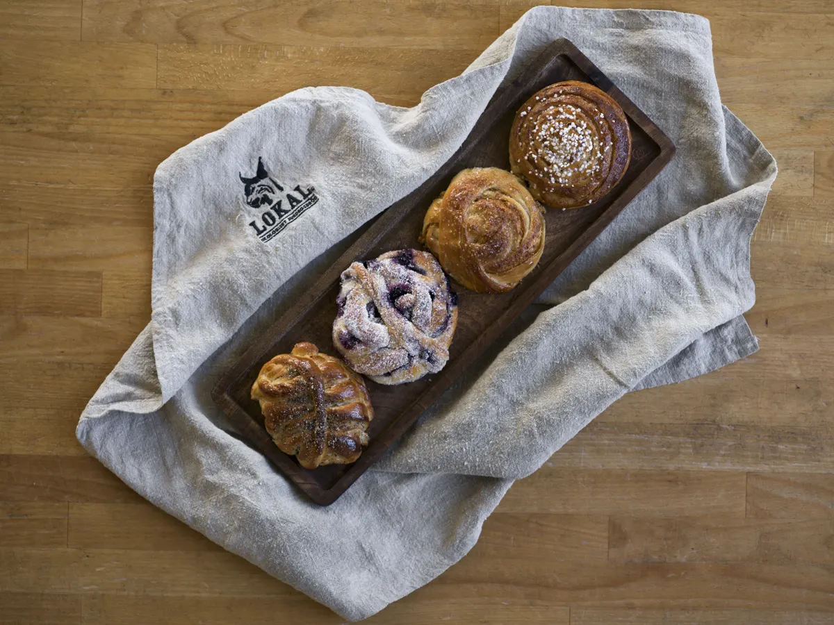 Fyra kanelbullar och en blåbärsbulle på ett träfat på en grå handduk med texten Lokal.