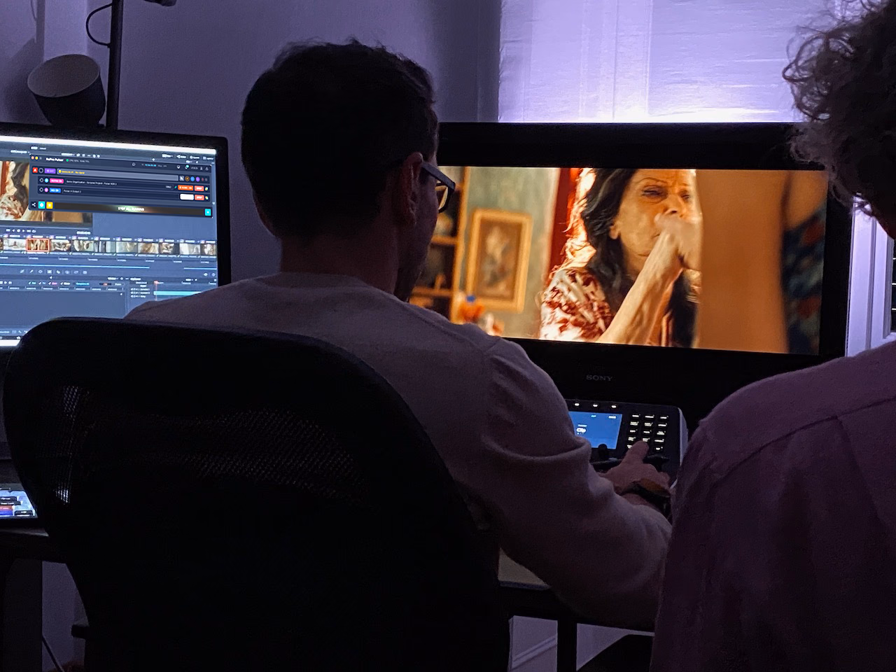 An editor in a grading suite with the edit timeline on one monitor and the programme picture on a larger display.