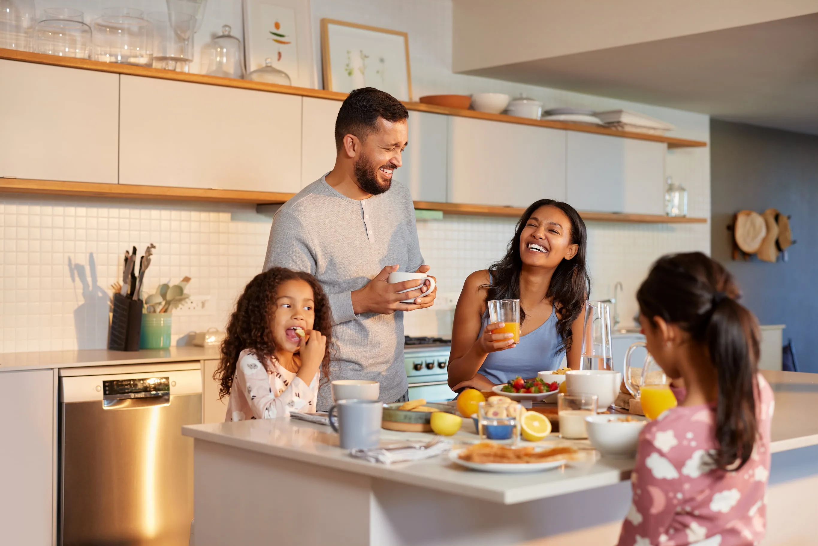 Una familia de cuatro personas se sienta y se para alrededor de una isla de cocina, desayunando y riendo juntos en una cocina moderna y luminosa.