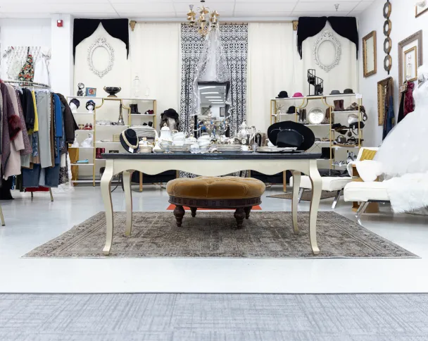 Elegant vintage store interior featuring chic clothing racks, decorative table with silverware, and a stylish round ottoman.