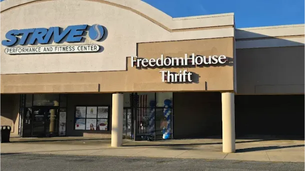 Exterior view of Freedom House Thrift store, adjacent to Strive Performance and Fitness Center, in a well-lit shopping area.