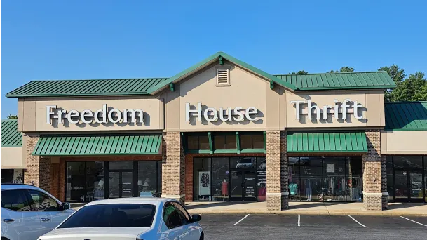 Exterior of Freedom House Thrift store with a green metal roof, large signage, and a clear blue sky in the backdrop.