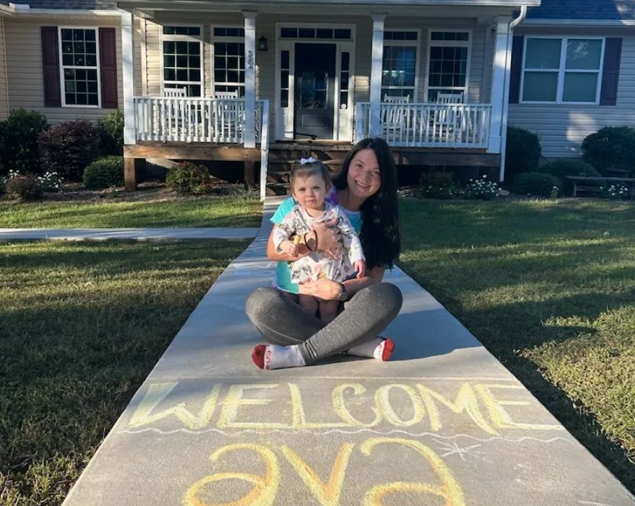 A woman sits on a sidewalk, holding a baby, with "WELCOME Ava" chalked on the concrete in front of a welcoming home.