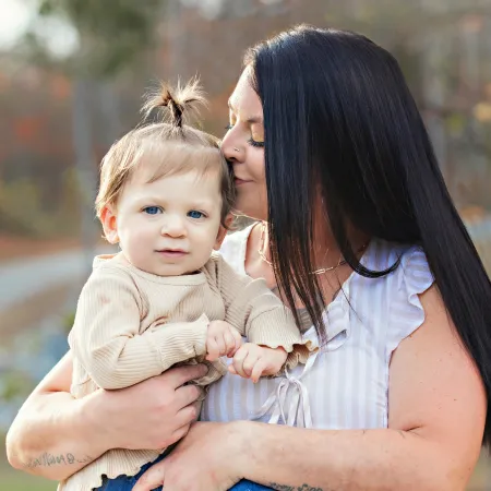 A woman with long dark hair holds a small child with a playful topknot, both outdoors in a soft-focused, natural setting.