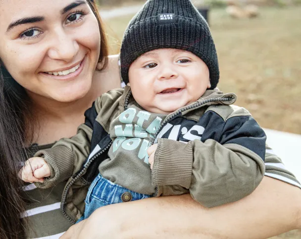 A child in a beanie and a hoodie is held by an adult. The background features a natural setting with soft-focus greenery.
