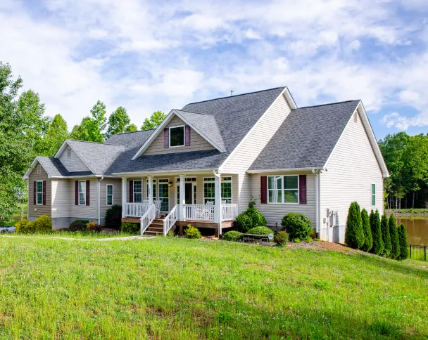 A spacious beige house with a gray roof, surrounded by green grass and trees, featuring a front porch and a tranquil pond in the background.