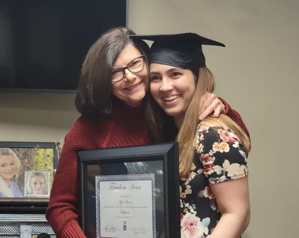 A graduate in a cap and gown embraces a loved one while holding a framed diploma, celebrating a significant achievement.