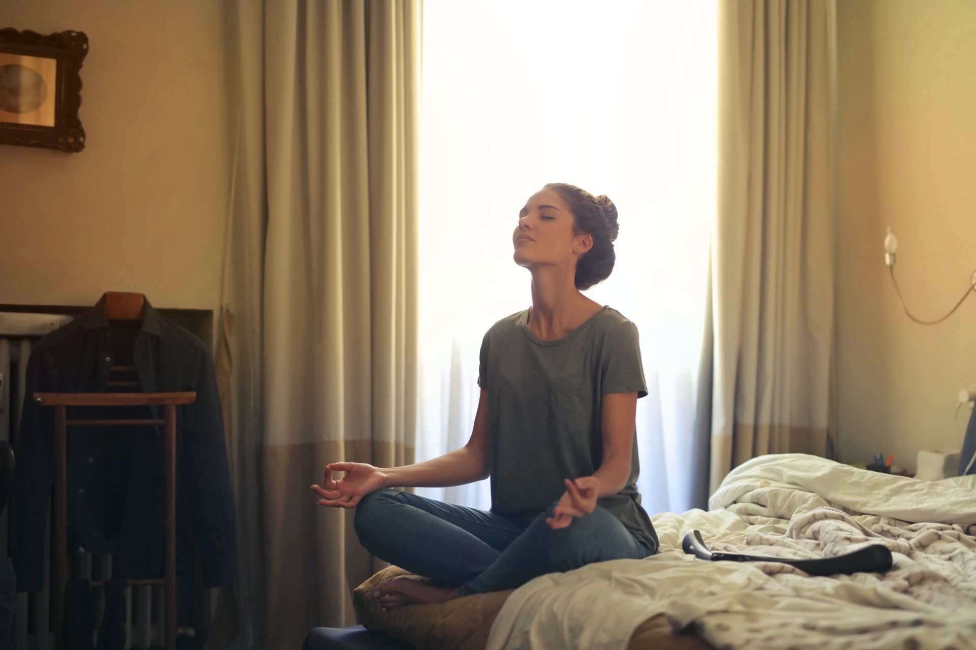Woman meditating in a cozy bedroom with natural light streaming through curtains, promoting relaxation and energy efficiency tips for summer comfort.