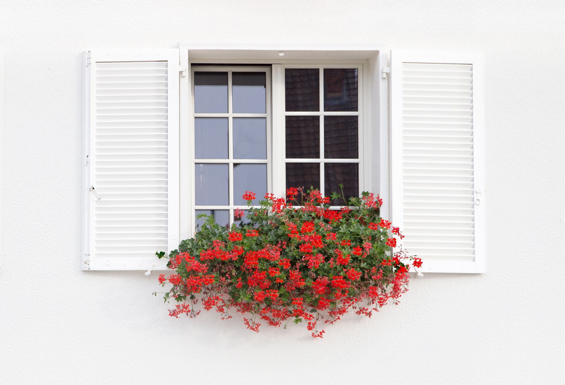 White window with open shutters and vibrant red flowers in a planter, symbolizing springtime and home maintenance for HVAC readiness.