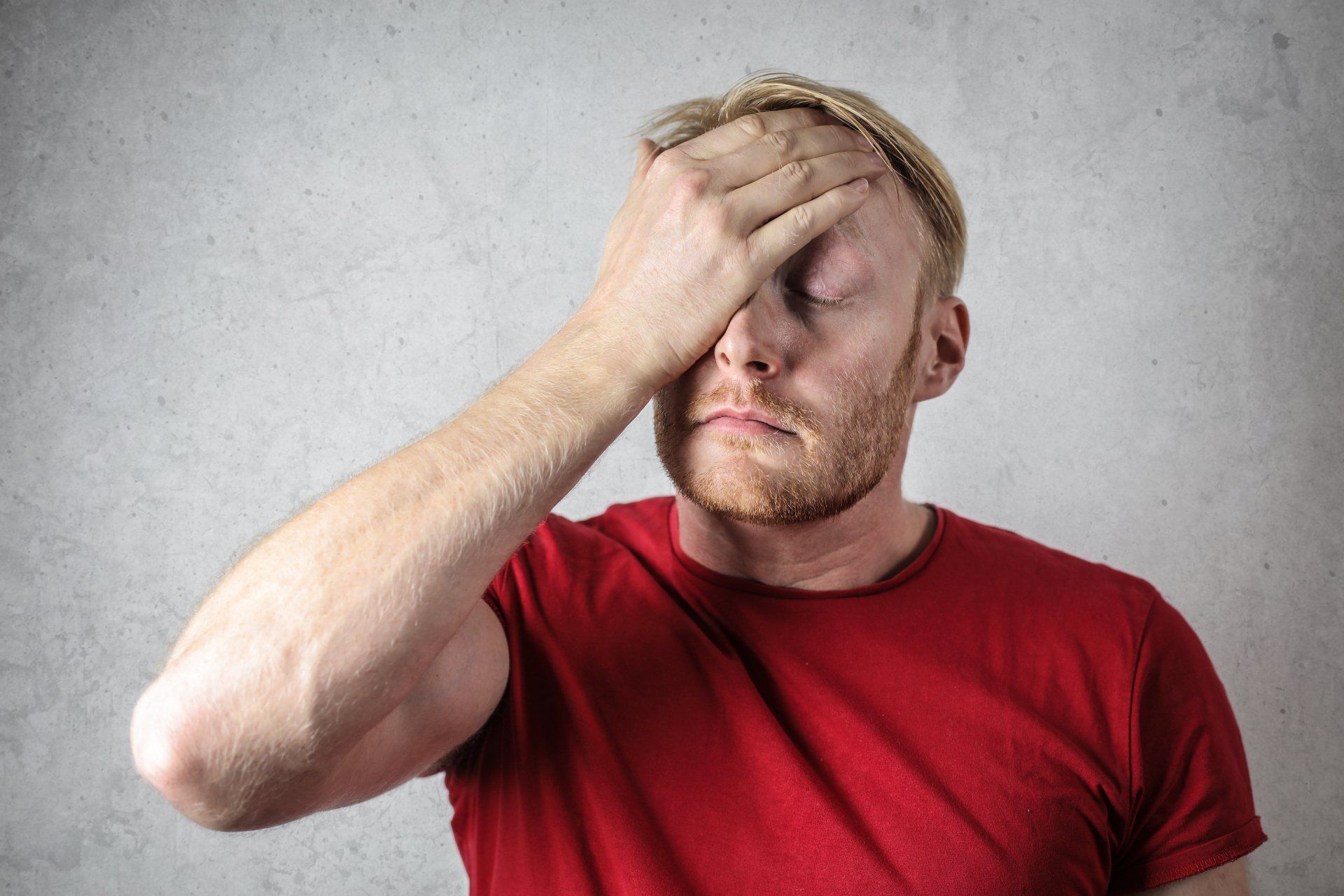 Man in a red shirt expressing frustration or concern, touching his forehead, against a textured gray background, relevant to HVAC system maintenance and repair discussions.