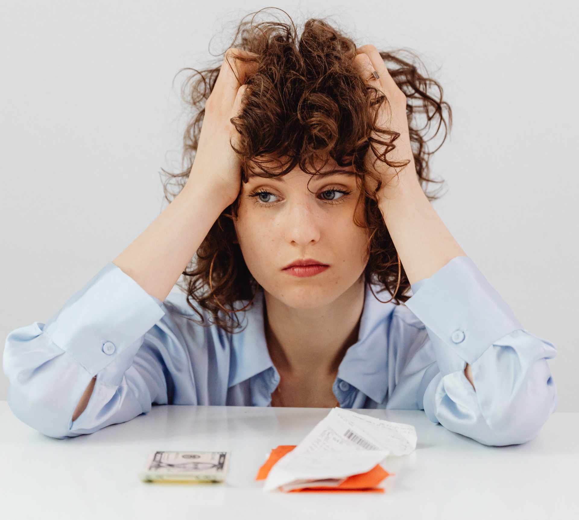 Woman with curly hair looking stressed, holding her head, with money and receipts on a table, reflecting concerns about home heating costs and energy efficiency.