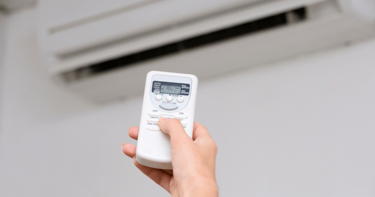 Hand holding air conditioner remote control, adjusting settings for optimal comfort and energy efficiency.