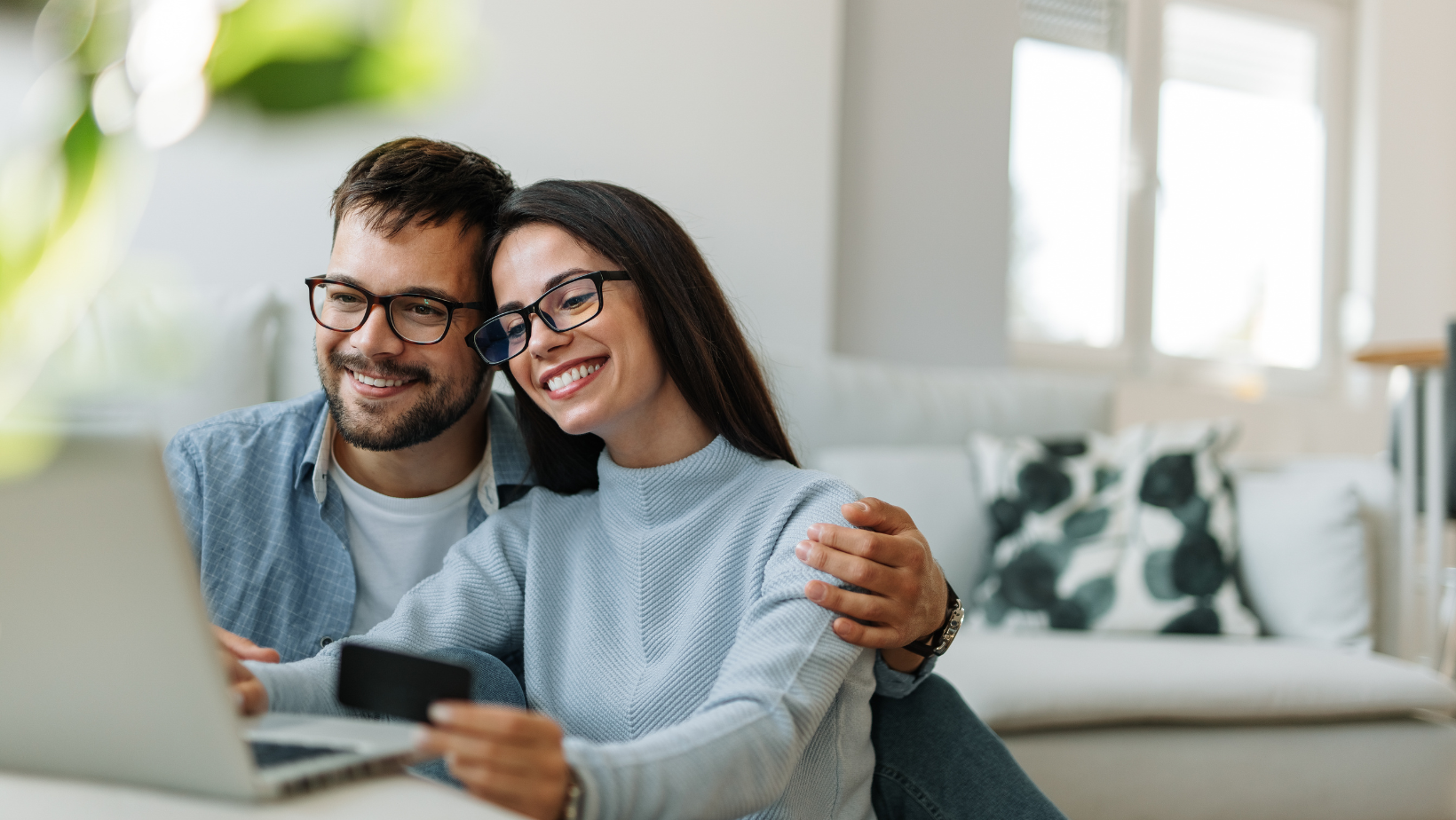 Couple smiling and using a laptop at home, demonstrating comfort and connection while considering HVAC services for seasonal air conditioning preparation.