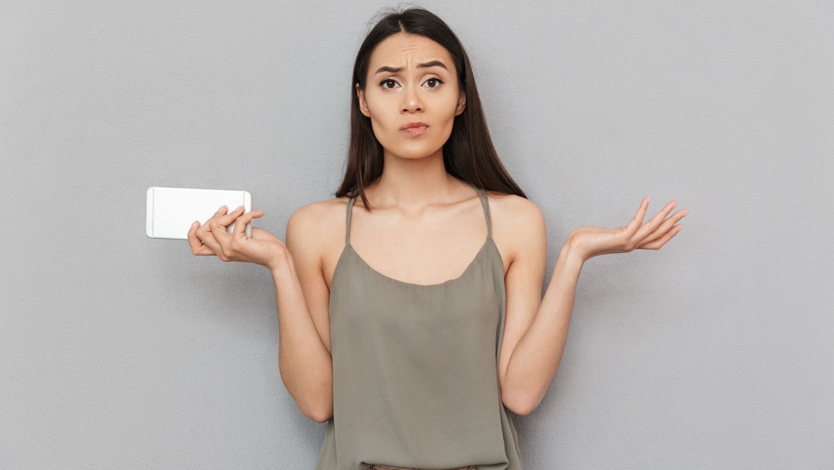 Confused woman holding a smartphone against a gray background, representing inquiries about HVAC maintenance and services.