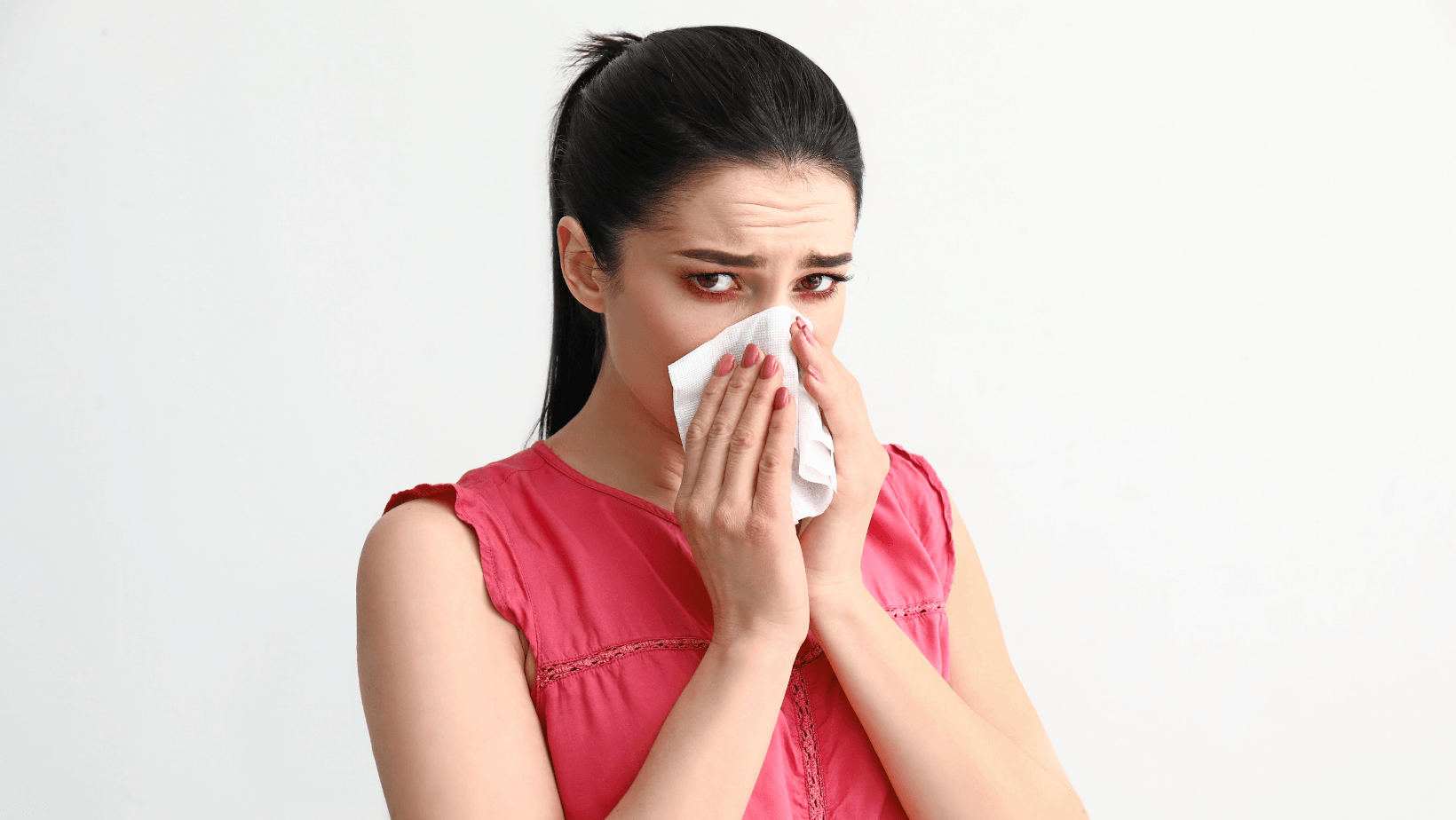 Woman in a pink shirt holding a tissue to her face, showing signs of allergy discomfort, emphasizing the importance of indoor air quality and HVAC maintenance for allergy relief.