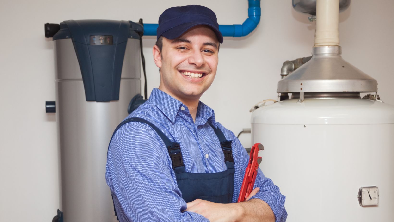 Smiling technician in blue shirt and cap holding red tools, standing next to a water heater, emphasizing HVAC and plumbing services.