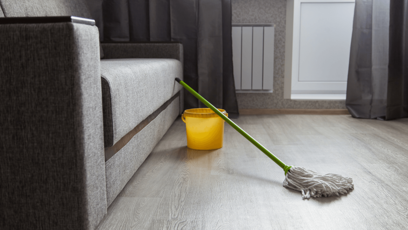 Mop and yellow bucket on wooden floor near a gray sofa, illustrating indoor cleaning process related to HVAC maintenance.
