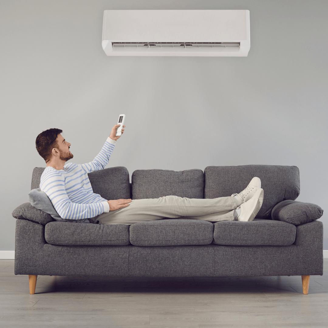 Man relaxing on a couch using a remote control for a ductless mini-split air conditioning unit, emphasizing HVAC comfort and air quality in a home setting.