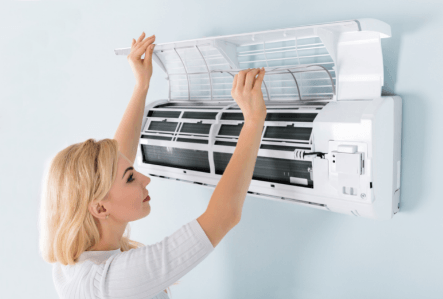 Woman performing maintenance on a wall-mounted air conditioning unit, highlighting the importance of air filter replacement for improved indoor air quality and HVAC system efficiency.
