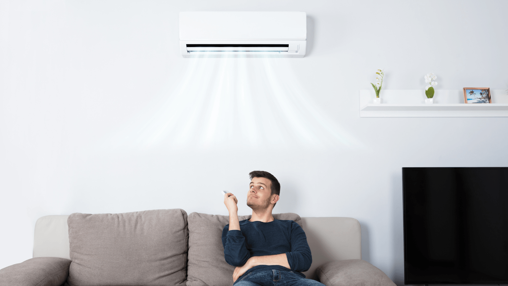 Man sitting on a couch using a remote control to adjust a wall-mounted ductless heat pump, emphasizing indoor air quality and HVAC services.