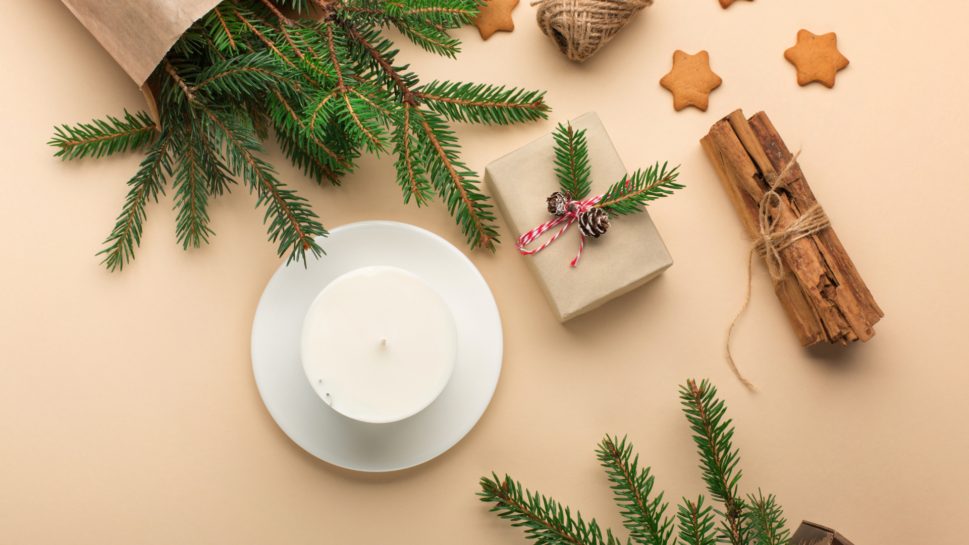 Eco-friendly home fragrance setup featuring a white candle on a plate, fresh pine branches, a wrapped gift with pinecone decoration, cinnamon sticks, and gingerbread cookies on a light background.