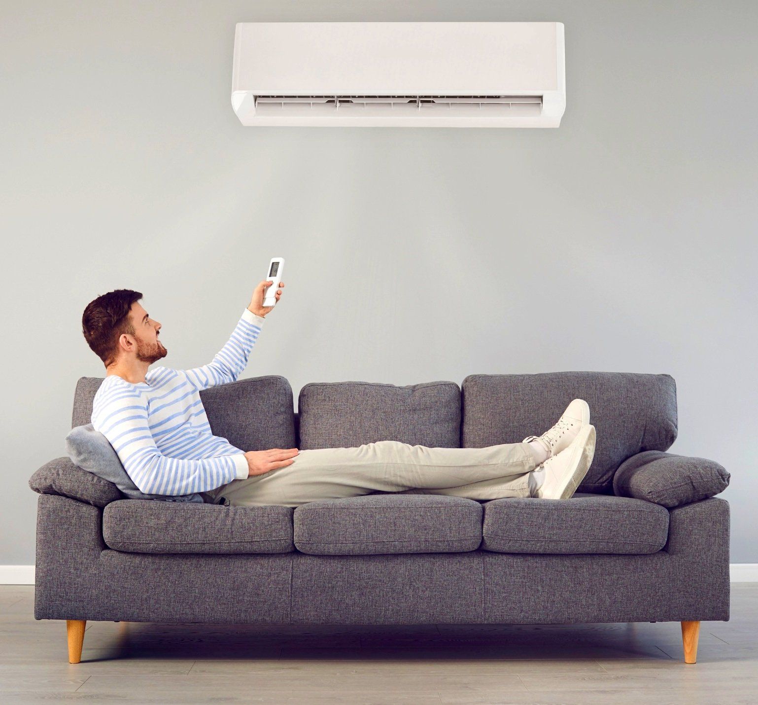 Man relaxing on a gray couch, using a remote control to adjust an air conditioning unit mounted on the wall, illustrating indoor comfort and temperature regulation.