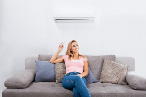 Woman sitting on a couch with a remote control, smiling at a wall-mounted HVAC unit, illustrating the importance of maintaining HVAC systems for comfort and efficiency.