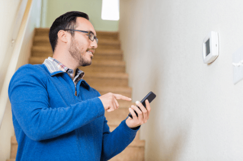 Man using smartphone to adjust smart thermostat on wall, illustrating energy-efficient HVAC management.